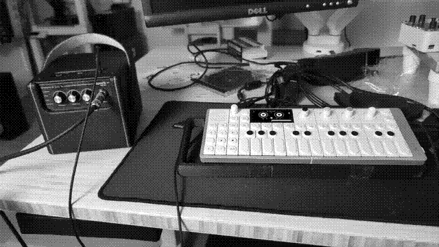 A dithered photo of a small guitar amplifier, no more than 6 or 7 inches along any dimension. It is plugged into a portable synthesizer and digital 4-track recorder, the OP-1 Field.