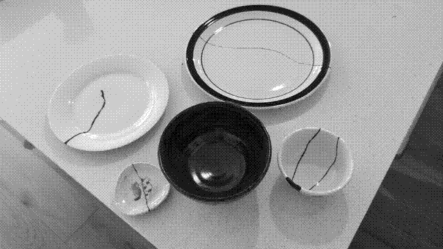 A grayscale photo from up higher. The two bowls in the previous photo are visible, along with three plates. The plates all have cracks down their middles. The smallest has two cats sitting together painted on it; the crack runs right between them.