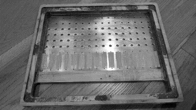 A back view of the mixer, open and without any electrical components inserted. The back of the aluminum faceplate is visible through a cavity that extends to within two centimeters of the edge of the wood in all directions, counting the one centimeter lip inset by 3.2 millimeters so the aluminum backplate sits flush with the wood when in place. Threaded inserts sit in dark mounds of putty made from sawdust and wood glue at the corners and midpoints of the edges. (They sit in putty because I routed the lip too deep and furthermore did not have an appropriately sized drillbit.) A three centimeter bit of wood, somewhere between 8 and 10 millimeters at its thickest point, divides the faceplate and miniplate. Bits of plastic vellum have been attached with double-sided tape over the slider slots and then split down the middle with a hobby knife, serving as dust covers.