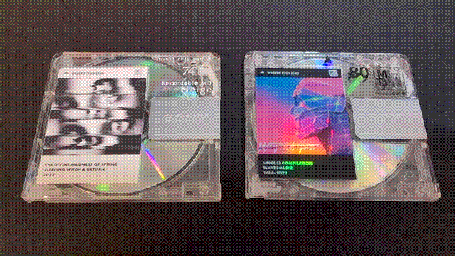 Two MiniDiscs. The one on the left has a black-and-white label for the Sleeping Witch & Saturn album "The Divine Madness of Spring" applied. It's a section of the album art, with the band name, album name, and release year (2022) in black-on-white Futura below, and the MiniDisc logo and "INSERT THIS END" above. The one on the right is formatted the same, but the artist is Waveshaper, the album is labeled "Singles Compilation," and the release year is "2014-2023." The artwork is the central part of the album art for "Hardware Passion" with the Waveshaper logo applied in the lower left corner. It's printed on holographic rainbow paper.
