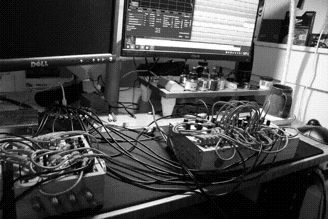 A grayscale photo of my desk. On my desk pad is my TAVELAN case, covered in patch cables and hooked up to an Expert Sleepers ES-9, itself without a case. A couple ribbon cables wired directly to a Meanwell RT-65B, visible just on the edge of the photo, power it all. Two flatscreen monitors are suspended by monitor arms above the desk, just behind the whole mess. One is off. The other shows Ardour running on my Steam Deck. Below this is a small desk shelf with multiple bottles of ink and a case containing a glass dip pen. A coffee mug sits atop an electric warming pad to the right, alongside a Kensington trackball.